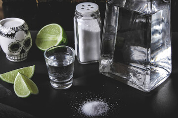 Tequila bottle and a salty jigger with lime and a Mexican skull next to them on a black surface