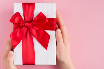 Holiday celebration concept. Top above overhead pov first person view photo of female hands holding white gift box with red ribbon isolated on pastel pink background with copyspace