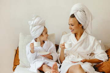 happy family mother and little daughter in bathrobe and towels rest and relax with a Cup of coffee in bed. - obrazy, fototapety, plakaty