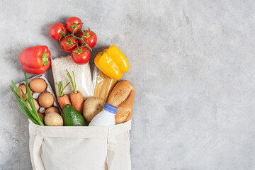 Cotton eco friendly shopping bag full of fresh organic vegetables and grocery products on grey table top view.