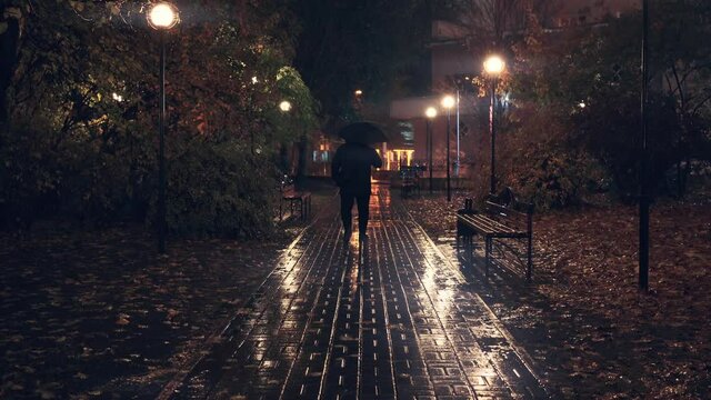 Man with umbrella walk alone on evening illuminated alley under rain in autumn night.