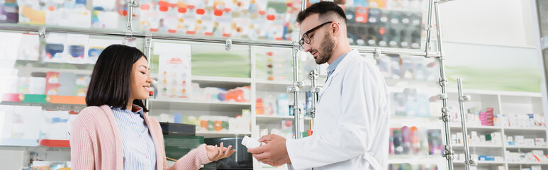 bearded pharmacist in eyeglasses and white coat giving bottle with pills to asian customer, banner
