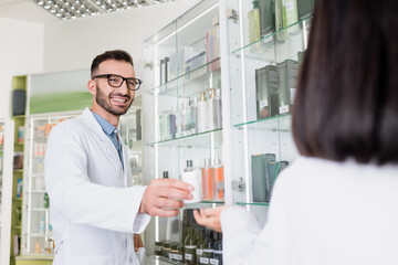 Obraz premium happy pharmacist in eyeglasses and white coat giving bottle with pills to brunette colleague on blurred foreground