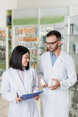 Obraz premium happy pharmacist in white coat holding bottles with medication near asian colleague in drugstore