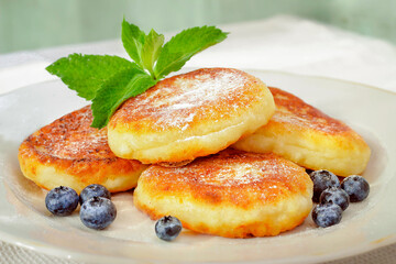 Cottage cheese pancakes, curd cakes, ricotta pancakes on a ceramic plate with powdered sugar, mint and fresh blueberries. Healthy and tasty morning breakfast. Close-up. Light background.