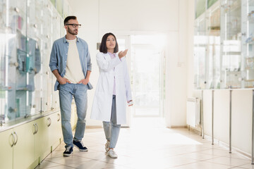 full length of asian pharmacist in white coat pointing with hand while walking with customer in drugstore