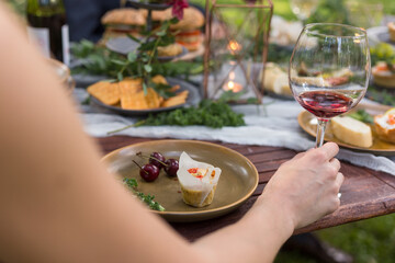 Female hand holding a glass of red wine at a rustic outdoor party. Plated savory muffin and cherries.