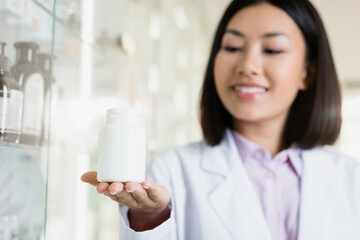 bottle with medication in hand of cheerful asian pharmacist on blurred background