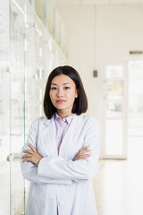 brunette asian pharmacist in white coat standing with crossed arms in drugstore