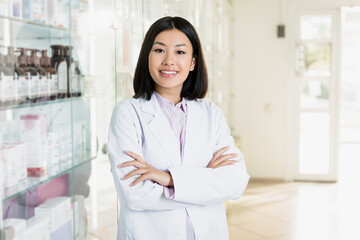 cheerful asian pharmacist in white coat standing with crossed arms in drugstore - obrazy, fototapety, plakaty