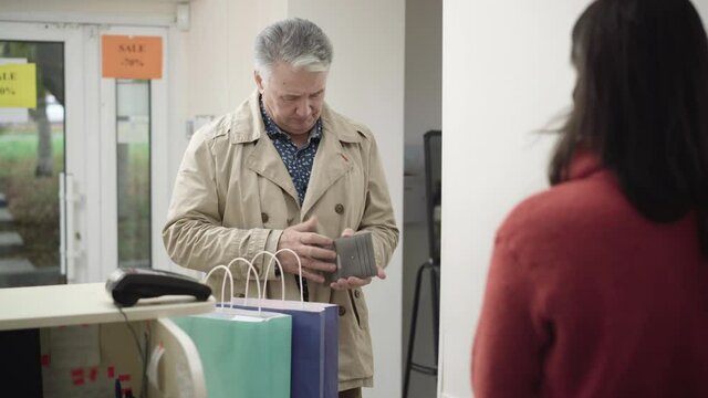 Senior grey-haired man not having enough money for payment in store. Portrait of embarrassed Caucasian male shopper looking in wallet, talking to shop assistant, and leaving.