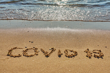 Covid 19 inscription on a tropical sandy beach with waves and foam on a background.