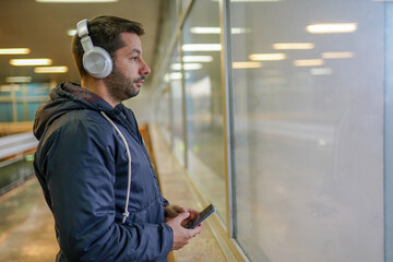 Young man with headphones standing on train station and looking through window while waiting train