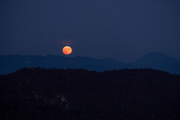 huge orange full moon rising behind a mountain ridge with layers of hills in the foreground creating a mystical atmosphere. glowing super moon rising into the night sky