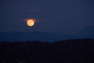 huge orange full moon rising behind a mountain ridge illuminating misty clouds creating a mystical atmosphere. glowing super moon rising into the night sky with layers of hills in the foreground.