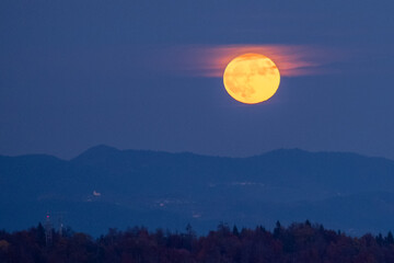 huge orange full moon rising behind a mountain ridge illuminating misty clouds creating a mystical atmosphere. glowing super moon rising into the night sky with layers of hills in the foreground.