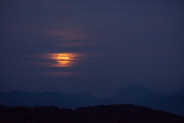 huge orange full moon rising behind a mountain ridge illuminating misty clouds creating a mystical atmosphere. glowing super moon rising into the night sky with layers of hills in the foreground.
