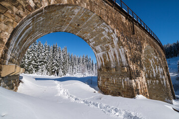 Stone viaduct (arch bridge) on railway through mountain snowy fir forest. Snow drifts  on wayside and hoarfrost on trees and electric line wires.