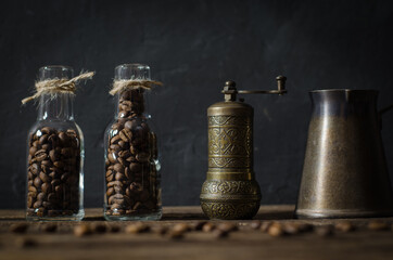 Stilllife with bottles filled by coffee beans, coffee grinder and coffee maker on the black background with scattered coffee beans