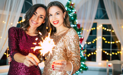 Two excited beautiful smiling girl friends in evening dresses are posing on the christmas tree background and having fun at the new year party
