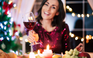 Excited gorgeous young happy woman in evening dress is holding a glass of wine and showing it to the camera while she sitting at christmas table