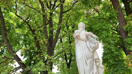 Footage of historical woman statue with trees in the background at famous 17th century garden called "Jardin Du Luxembourg" in Paris. It is a sunny summer day. Camera pans right.