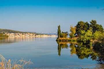Fototapeta premium Koutavos Lagoon's serene waters mirror the charming town of Argostoli under a vast blue sky. Lush greenery frames the tranquil scene, offering a peaceful escape in the heart of Kefalonia, Greece.