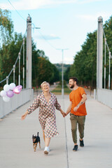 Cheerful lovely couple walking happy on bridge with their dog and pink balloons smiling