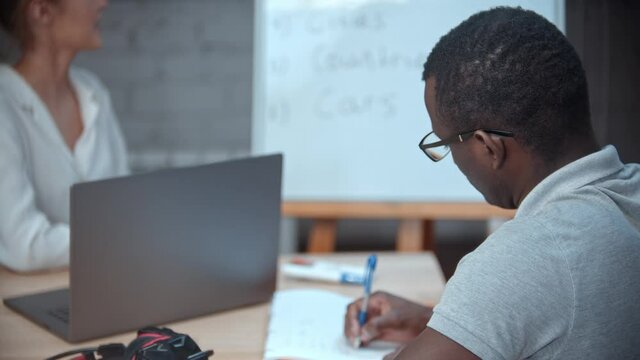 Woman teacher giving a black man an english lesson - pointing at the board with themes for the first lesson and man writes it in his notebook