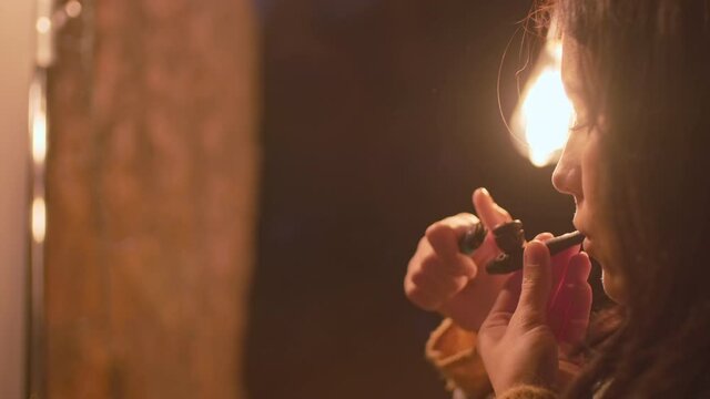 Close up of young beautiful brunette holding pipe and lighter in hands smoking slowly enjoying flavor and aroma