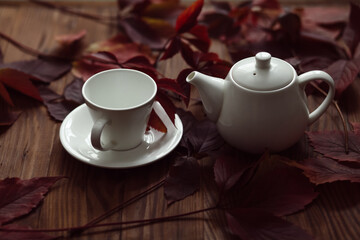 Close-up of a white cup and a teapot on a table with a background of autumn leaves. Autumn rustic still life of the tea ceremony.