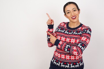 Young beautiful Arab woman wearing Christmas sweater, against white background indicating finger empty space showing best low prices, looking at the camera