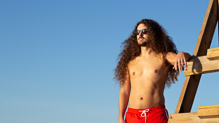 Closeup shot of an attractive young man with long curly hair leaning on a lifeguard chair