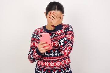 Young beautiful Arab woman wearing Christmas sweater against white wall,  looking at smart phone feeling sad holding hand on face.