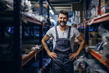 Smiling tattooed bearded worker in overalls standing in storage with hands on hips.