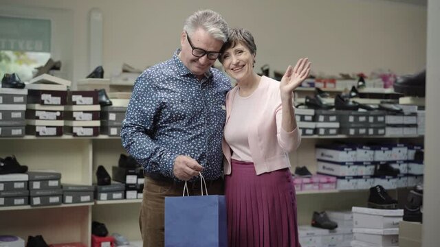 Portrait Of Loving Senior Caucasian Couple Posing In Shoe Store During Black Friday Sales. Happy Positive Man And Woman Waving At Camera And Smiling As Standing With Shopping Bags In Shop.