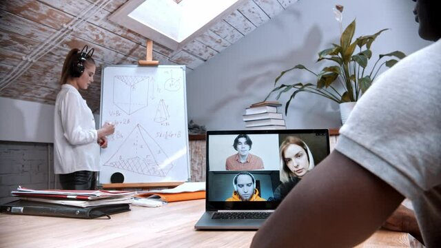 An online maths lesson - woman standing by the board and her students on the screen listening to her