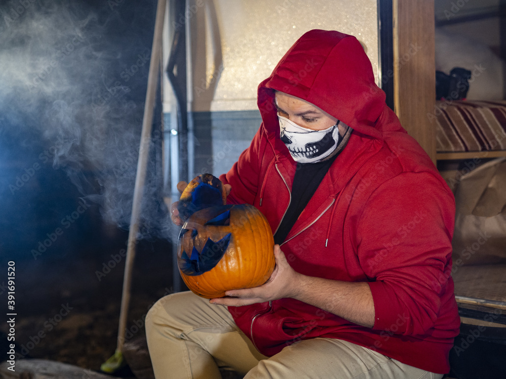 Sticker Person in a red hoodie and a skull facemask shading a carved Halloween pumpkin