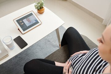 Pregnant young woman touching her belly when making video call to her doctor