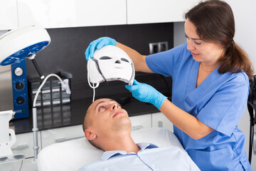 Young man receiving facial light therapy in aesthetic medicine office. Female cosmetologist putting...