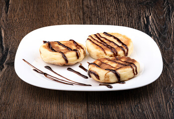 Three cheese pancakes on a white plate on a wooden background. Cheese pancakes decorated with chocolate topping.