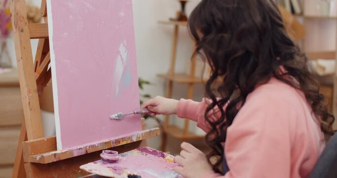 Backside view of brunnete girl with long wavy hair creating picture. Young lady sitting in front of molbert and holding palette while drawing with spatula and acrylic paints. Concept of art.