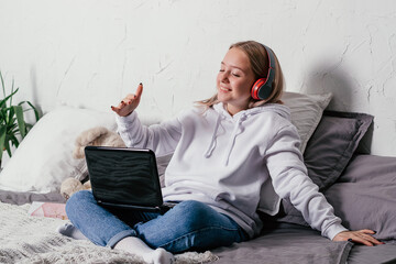 Video communication.Teenage girl with wireless headphones sitting   on the bed and communicating via video chat,zoom using laptop.Communication and education in the coronavirus pandemic.