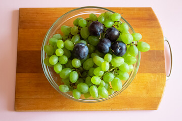 Green grapes and blue plums in a transparent glass dish on a wooden surface. Top view. Fruit diet. Summer fruit