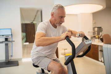 Naklejka na ściany i meble Grey-haired man sitting on a treadmill looking concentrated and determined
