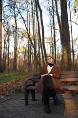 Fototapeta premium A charming girl with blonde hair , wearing a brown wool coat, is sitting on a wooden bench in an alley in an autumn Park.