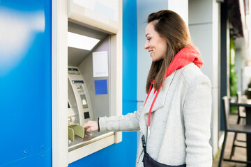 Young brunette woman inserting credit card, withdrawing money at outdoor ATM.