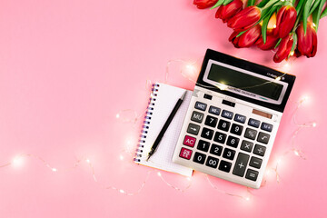 Large silver calculator, black pen, spiral notepad, lights garland and tulips bouquet lays on a pink background. Concept of business holiday and congratulating women on women's day at the workplace