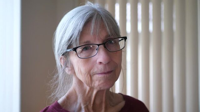 An old caucasian woman with aging face wrinkles gray hair and glasses looking at camera hopeful, calm and happy.
