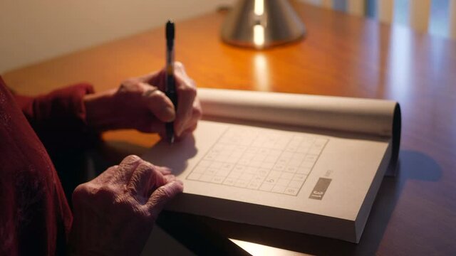 Middle Aged Caucasian Woman With Aging Hands Writing With A Pen In A Number Puzzle Book Solving For Mind Acuity To Stay Mentally Adept And Healthy.